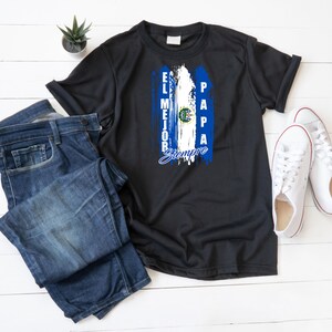 Peut inclure: T-shirt noir avec un motif du drapeau salvadorien. Le motif comprend des rayures verticales bleues et blanches avec le texte "El Mejor Papa Siempre". Le t-shirt est présenté avec un jean bleu et des baskets blanches.