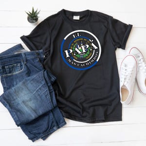 Peut inclure: T-shirt noir avec un motif circulaire portant l'inscription "EL PAPÁ" et les mots "EL SALVADOR EN LA REPUBLICA CENTRAL" en blanc et bleu. Le t-shirt est associé à un jean bleu et à des baskets blanches.