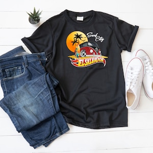 Peut inclure: T-shirt noir avec un motif représentant un van vintage rouge et noir, une planche de surf et un coucher de soleil. Le texte "Surf City El Salvador" est affiché au-dessus et en dessous du van. Un jean bleu et des baskets blanches sont également présents.