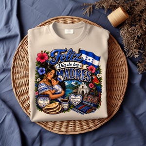 May include: Beige sweatshirt featuring a colorful graphic celebrating Mother's Day. The design includes the text "Feliz Día de las Madres" with illustrations of a mother and child, flowers, a parrot, and the Honduran flag.