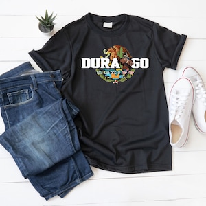 May include: Black t-shirt featuring the word "DURANGO" in white, above a colourful Mexican emblem. The shirt is displayed with blue jeans and white trainers on a white wooden surface.