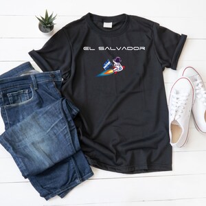 Peut inclure: T-shirt noir avec du texte blanc qui dit "EL SALVADOR" et un astronaute de dessin animé tenant un drapeau salvadorien sur une fusée.