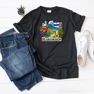 Peut inclure: T-shirt noir avec un motif coloré d'El Salvador, comprenant un perroquet, un volcan, un drapeau et du texte. Le t-shirt est associé à un jean bleu et des baskets blanches, pour un look décontracté.