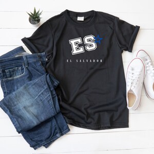 Peut inclure: T-shirt noir avec inscription blanche "ES" surmontée d'une étoile bleue et "EL SALVADOR" en dessous. Le t-shirt est posé à plat sur une surface en bois blanc avec un jean bleu et des baskets blanches.
