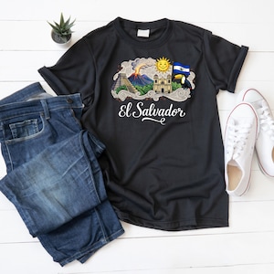 Peut inclure: T-shirt noir avec un graphisme coloré des monuments du Salvador, dont un volcan, une église et un drapeau. Le texte "El Salvador" est écrit en blanc. Le t-shirt est présenté avec un jean bleu et des baskets blanches.