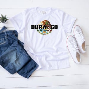 Op de afbeelding: Wit T-shirt met het woord "DURANGO" in zwart boven een kleurrijk embleem en het woord "MEXICO". Het shirt wordt getoond met een blauwe jeans en witte sneakers, tegen een witte houten achtergrond.
