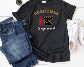 May include: Black t-shirt with the word "GUATEMALA" in gold lettering and a rooster logo. The shirt also features the text "Gallo" and "la mejor cerveza!" in red and white. The shirt is paired with blue jeans and white sneakers.