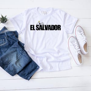 May include: A white t-shirt with the text "EL SALVADOR" in black, and a cartoon character. The shirt is displayed with blue jeans and white trainers. A small plant is on a white wooden surface.
