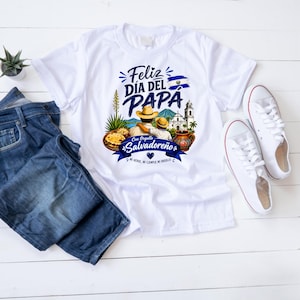 May include: White t-shirt with a graphic design celebrating Father's Day in El Salvador. The design includes text in Spanish, illustrations of food, a church, and the Salvadoran flag. The shirt is paired with blue jeans and white sneakers.