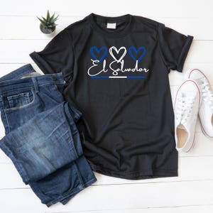 Peut inclure: T-shirt noir avec l'inscription "El Salvador" en écriture blanche, des cœurs bleus et blancs, et une bande horizontale bleue et blanche. Le t-shirt est associé à un jean bleu et des baskets blanches.