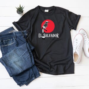 Peut inclure: T-shirt noir avec un cercle rouge et un motif d'oiseau blanc. Le texte "El Salvador" est imprimé en blanc sous le motif.