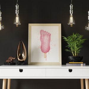 Puede incluir: Una pintura al agua enmarcada de una paleta de helado rosa en forma de pie con pintura rosa y blanca goteando sobre un fondo blanco. La pintura est&aacute; colgada en una pared blanca sobre un escritorio blanco con una tapa de madera y dos cajones. Hay cuatro bombillas colgantes sobre el escritorio, y una peque&ntilde;a planta en maceta y dos objetos decorativos en el escritorio.