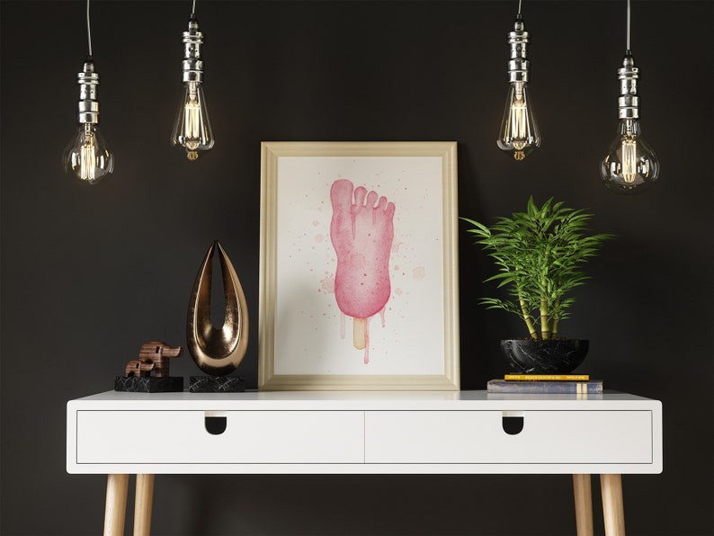 Puede incluir: Una pintura al agua enmarcada de una paleta de helado rosa en forma de pie con pintura rosa y blanca goteando sobre un fondo blanco. La pintura est&aacute; colgada en una pared blanca sobre un escritorio blanco con una tapa de madera y dos cajones. Hay cuatro bombillas colgantes sobre el escritorio, y una peque&ntilde;a planta en maceta y dos objetos decorativos en el escritorio.