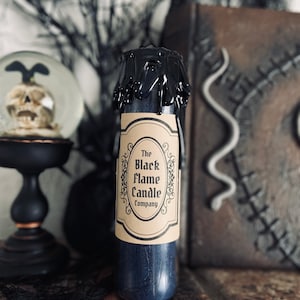 May include: A black candle with a label that reads "The Black Flame Candle Company". The candle is dripping black wax and is sitting on a brick surface.