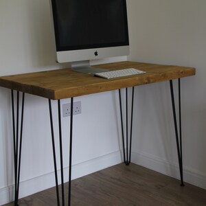 May include: A wooden desk with black hairpin legs. The desk has a light brown wood top and is sitting on a wood floor. A computer monitor and keyboard are on the desk.