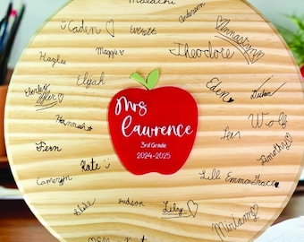 May include: A round wooden teacher appreciation plaque with student signatures. A red apple with the text "Mrs. Lawrence 3rd Grade 2024-2025" is in the center. The wooden plaque has a light wood grain.