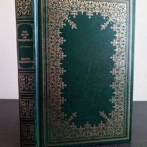 May include: A hardcover book titled "The Vicar of Wakefield" by Oliver Goldsmith. The book has a dark green cover with gold decorative filigree and lettering. The spine also features gold accents and the title.