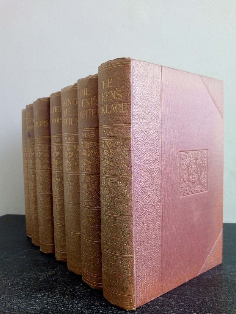 Puede incluir: Una colecci&oacute;n de libros antiguos con cubiertas marrones y letras doradas. Los t&iacute;tulos incluyen "El collar de la reina" y "La hija del rey". Los libros est&aacute;n dispuestos en fila, con un libro rosa en el lado derecho.