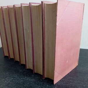 Puede incluir: Una fila de libros de tapa dura con cubiertas de color rosa empolvado y lomos burdeos. Las p&aacute;ginas est&aacute;n envejecidas, con un color marr&oacute;n claro. Los libros est&aacute;n dispuestos en diagonal, lo que sugiere que est&aacute;n en una estanter&iacute;a o mesa.