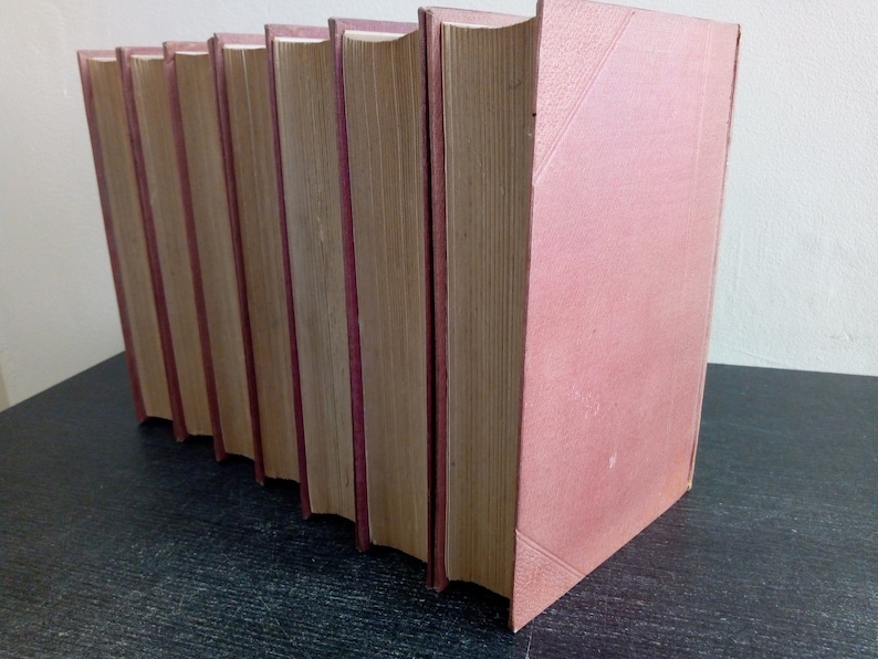 Puede incluir: Una fila de libros de tapa dura con cubiertas de color rosa empolvado y lomos burdeos. Las p&aacute;ginas est&aacute;n envejecidas, con un color marr&oacute;n claro. Los libros est&aacute;n dispuestos en diagonal, lo que sugiere que est&aacute;n en una estanter&iacute;a o mesa.