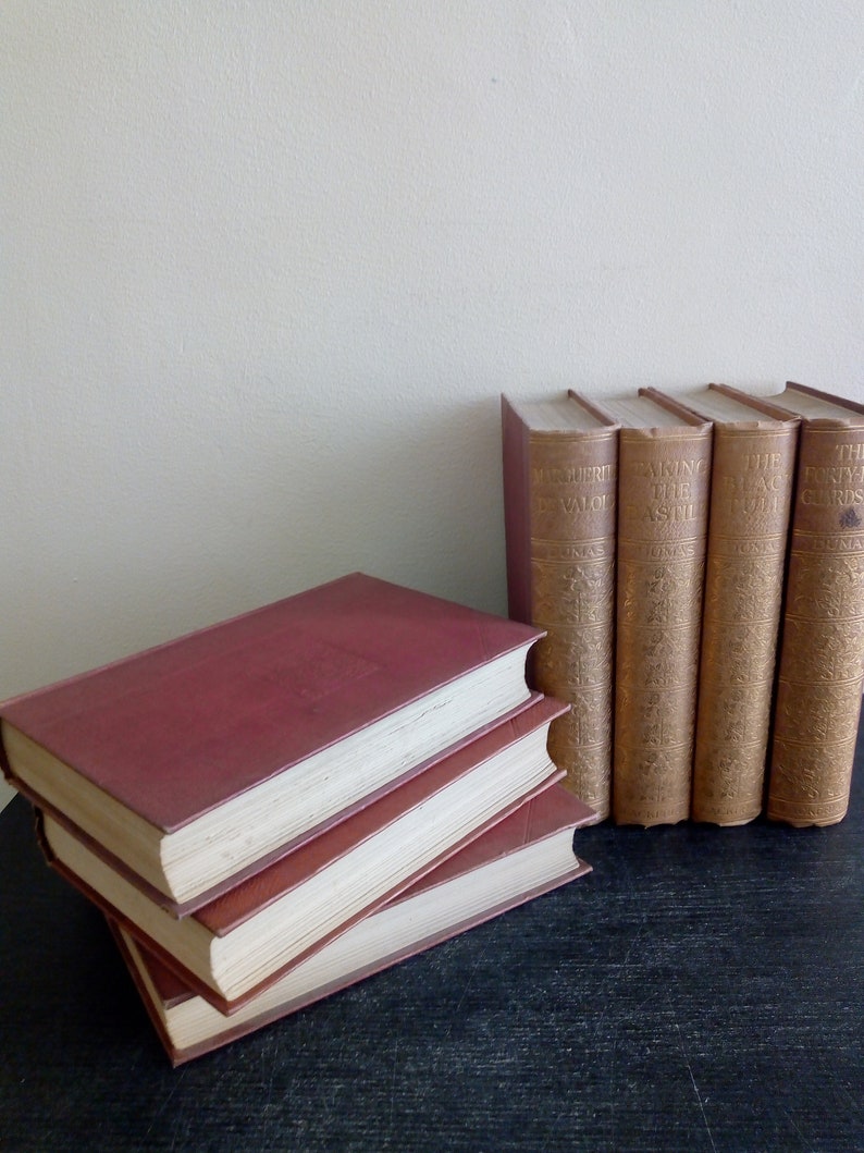 Puede incluir: Una colecci&oacute;n de libros antiguos, con una pila de tres vol&uacute;menes burdeos y una fila de cinco libros de pie. Los libros tienen letras doradas y cubiertas texturizadas envejecidas, con p&aacute;ginas blanquecinas.