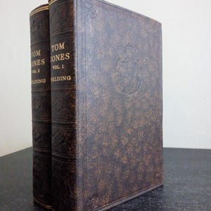Puede incluir: Se muestran dos libros antiguos, "Tom Jones" Volumen 1 y Volumen 2. Los libros tienen cubiertas de cuero marrón oscuro con letras y detalles dorados. Las cubiertas presentan diseños en relieve y los libros están sobre una superficie oscura.