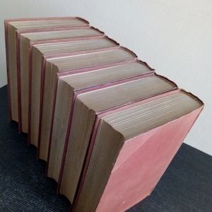 Puede incluir: Colecci&oacute;n de libros antiguos con cubiertas burdeos, dispuestos en diagonal descendente. Las p&aacute;ginas est&aacute;n envejecidas y las lomos muestran desgaste. Los libros est&aacute;n apilados sobre una superficie oscura.