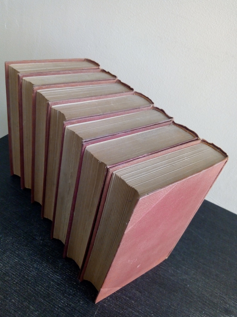 Puede incluir: Colecci&oacute;n de libros antiguos con cubiertas burdeos, dispuestos en diagonal descendente. Las p&aacute;ginas est&aacute;n envejecidas y las lomos muestran desgaste. Los libros est&aacute;n apilados sobre una superficie oscura.