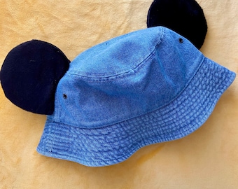 May include: Blue denim bucket hat with two large, black, plush ears attached. The hat has a classic bucket hat design with a slightly faded appearance. The ears are round and fluffy, adding a playful touch to the hat's style.
