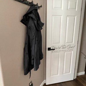 May include: A black coat hangs on a black metal coat rack with five hooks. The coat rack is mounted on a light brown wall. A white door with a black door handle is to the right of the coat rack. The door has a black vinyl sticker that says "You're our Guest Make Yourself at Home".