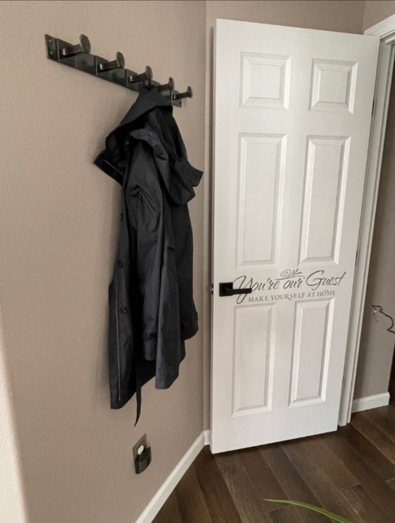May include: A black coat hangs on a black metal coat rack with five hooks. The coat rack is mounted on a light brown wall. A white door with a black door handle is to the right of the coat rack. The door has a black vinyl sticker that says "You're our Guest Make Yourself at Home".