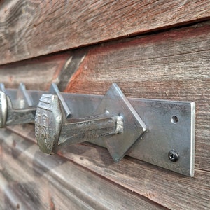 May include: A metal coat rack with five large nail hooks mounted on a rectangular metal plate. The rack is attached to a wooden wall with a black screw.