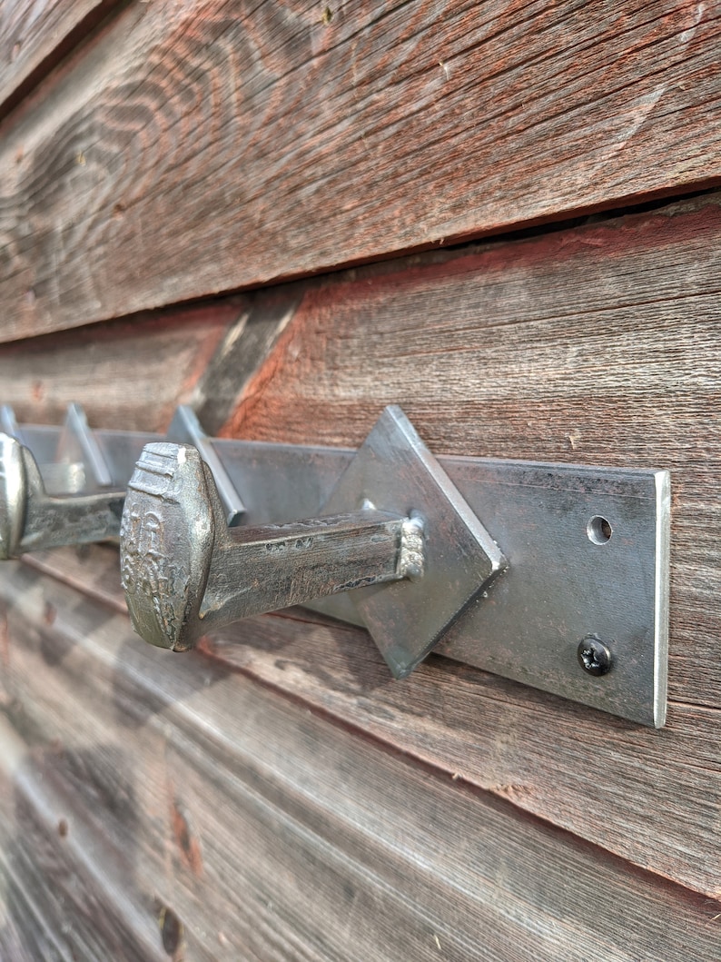 May include: A metal coat rack with five large nail hooks mounted on a rectangular metal plate. The rack is attached to a wooden wall with a black screw.