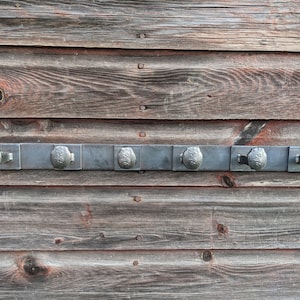 May include: A metal coat rack with six hooks, mounted on a wooden wall. The hooks are made of metal and have a rounded shape. The wall is made of wood and has a natural finish.