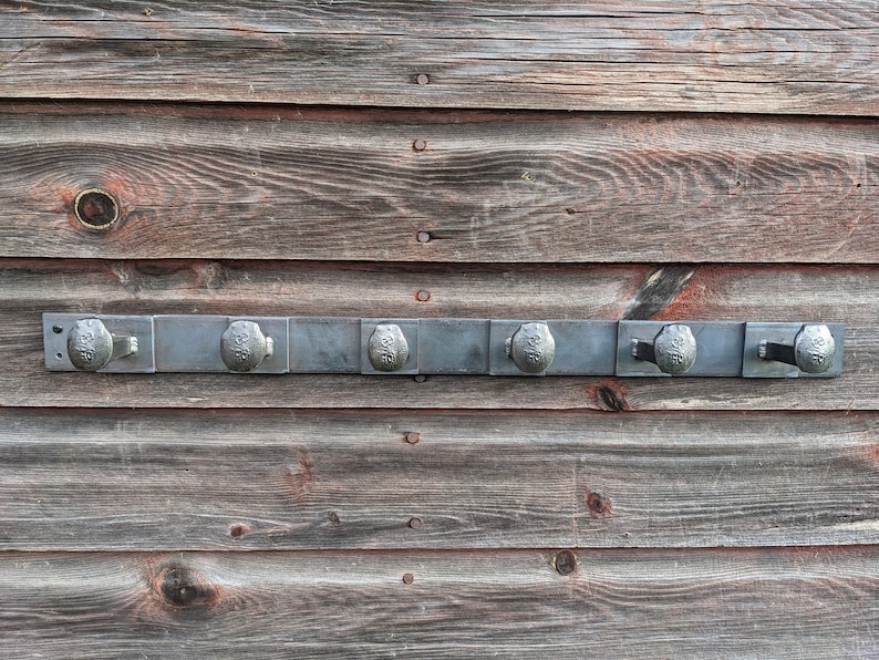 May include: A metal coat rack with six hooks, mounted on a wooden wall. The hooks are made of metal and have a rounded shape. The wall is made of wood and has a natural finish.