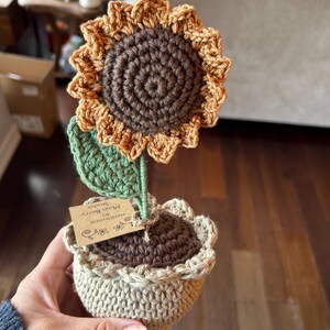 May include: A crocheted sunflower in a tan pot with a brown center and golden-yellow petals. A green leaf is attached to the stem. A small brown paper tag is tied to the stem with the text "HANDMADE BY Mojo Berry Studio" and a flower icon.