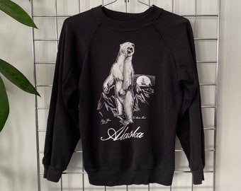 May include: Black sweatshirt featuring a graphic of a polar bear standing on its hind legs, with the word "Alaska" in cursive script. The design includes a mountain range and a ship. The sweatshirt is hanging on a metal grid.