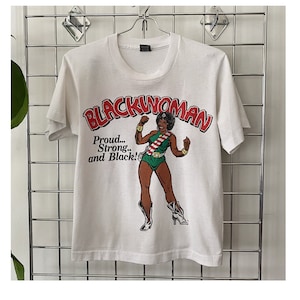 K&ouml;nnte beinhalten: Wei&szlig;es T-Shirt mit einem Grafikdruck einer schwarzen Frau in einem gr&uuml;n-roten Kost&uuml;m mit dem Text "Blackwoman" in roten Buchstaben und "Proud... Strong... and Black!" in schwarzen Buchstaben.
