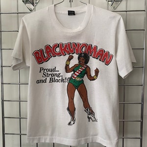 K&ouml;nnte beinhalten: Wei&szlig;es T-Shirt mit einem Grafikdruck einer schwarzen Frau in einem gr&uuml;n-wei&szlig;en Kost&uuml;m mit dem Text "Blackwoman" in rot-wei&szlig;en Buchstaben und dem Text "Proud... Strong... and Black!" in schwarzen Buchstaben.