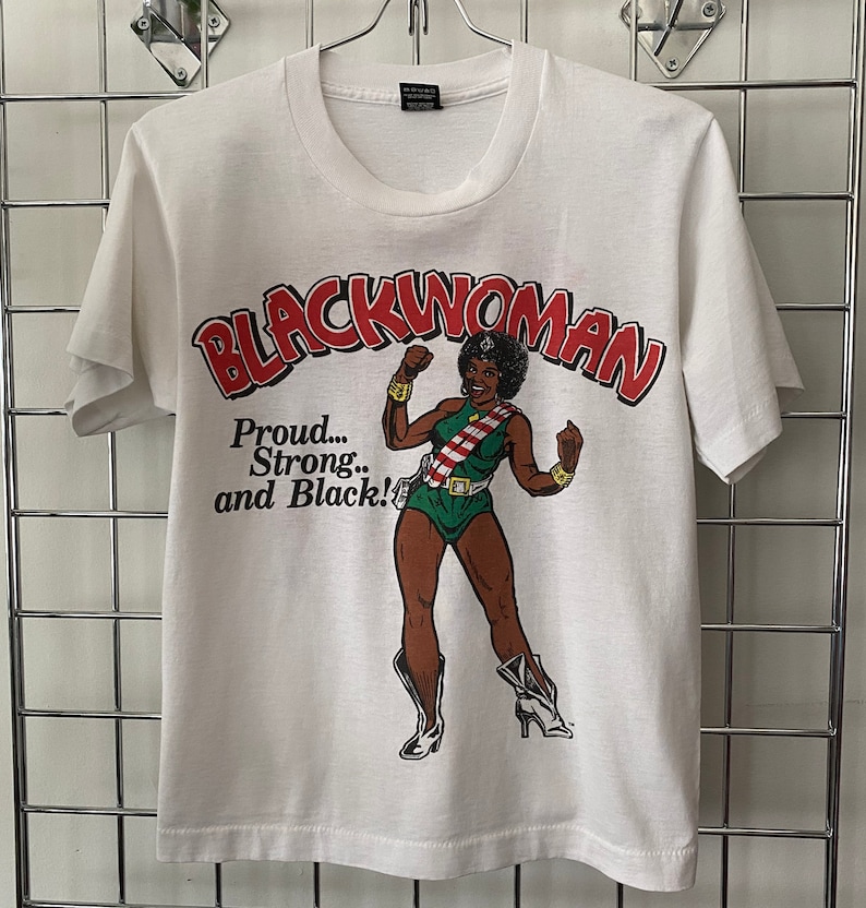 K&ouml;nnte beinhalten: Wei&szlig;es T-Shirt mit einem Grafikdruck einer schwarzen Frau in einem gr&uuml;n-wei&szlig;en Kost&uuml;m mit dem Text "Blackwoman" in rot-wei&szlig;en Buchstaben und dem Text "Proud... Strong... and Black!" in schwarzen Buchstaben.