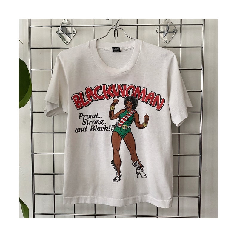 K&ouml;nnte beinhalten: Wei&szlig;es T-Shirt mit einem Grafikdruck einer schwarzen Frau in einem gr&uuml;n-roten Kost&uuml;m mit dem Text "Blackwoman" in roten Buchstaben und "Proud... Strong... and Black!" in schwarzen Buchstaben.
