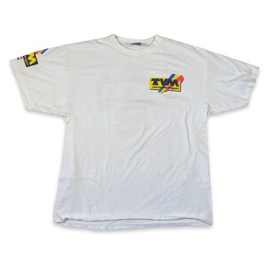 May include: White t-shirt with a yellow, blue, red, and black logo that says "TVM Verzekeringen" on the front. The logo is on a diagonal with a yellow background. The shirt has a blue, red, and yellow stripe on the left sleeve.