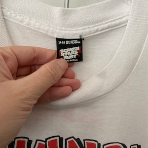 K&ouml;nnte beinhalten: Wei&szlig;es T-Shirt mit einem roten und schwarzen Grafikdruck des Wortes "SKWOK" im Cartoon-Stil. Das Etikett auf dem Shirt lautet "Screen Stars Best" und "50% Baumwolle 50% Polyester" und "Hergestellt in den USA."