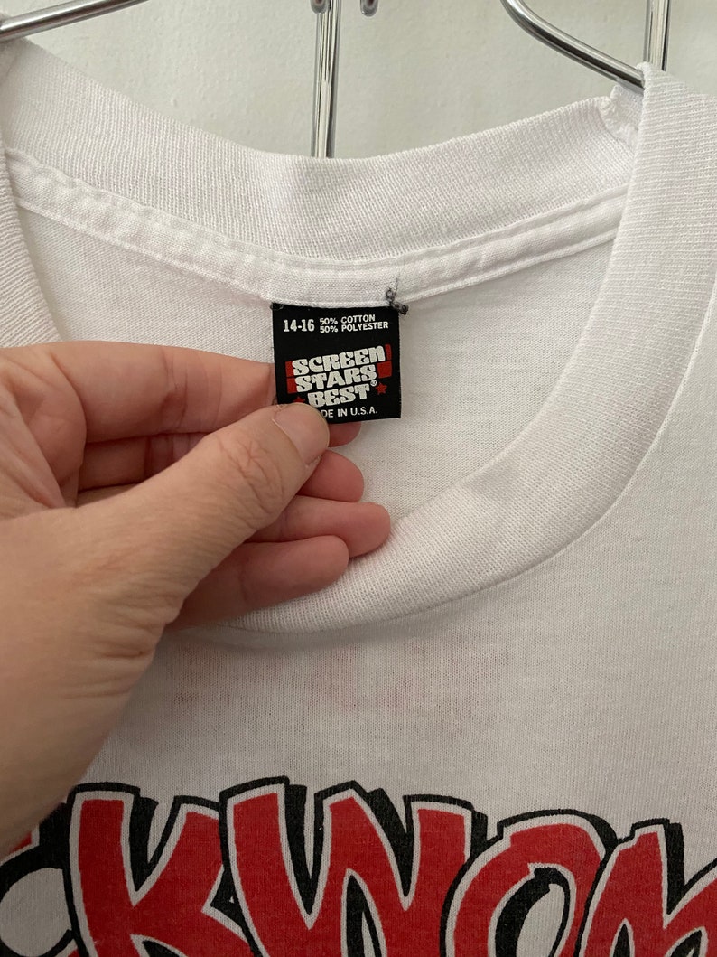 K&ouml;nnte beinhalten: Wei&szlig;es T-Shirt mit einem roten und schwarzen Grafikdruck des Wortes "SKWOK" im Cartoon-Stil. Das Etikett auf dem Shirt lautet "Screen Stars Best" und "50% Baumwolle 50% Polyester" und "Hergestellt in den USA."