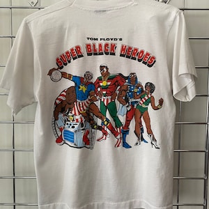 K&ouml;nnte beinhalten: Wei&szlig;es T-Shirt mit einem Aufdruck einer Gruppe schwarzer Superhelden. Der Aufdruck lautet "Tom Floyd's Super Black Heroes".