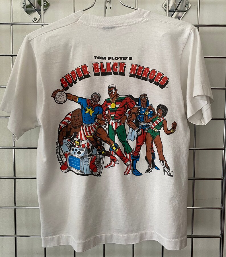 K&ouml;nnte beinhalten: Wei&szlig;es T-Shirt mit einem Aufdruck einer Gruppe schwarzer Superhelden. Der Aufdruck lautet "Tom Floyd's Super Black Heroes".