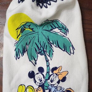 May include: White kitchen towel with a blue crocheted hanging loop and a button. The towel features a cartoon image of Mickey Mouse and Minnie Mouse under a palm tree with a yellow sun.