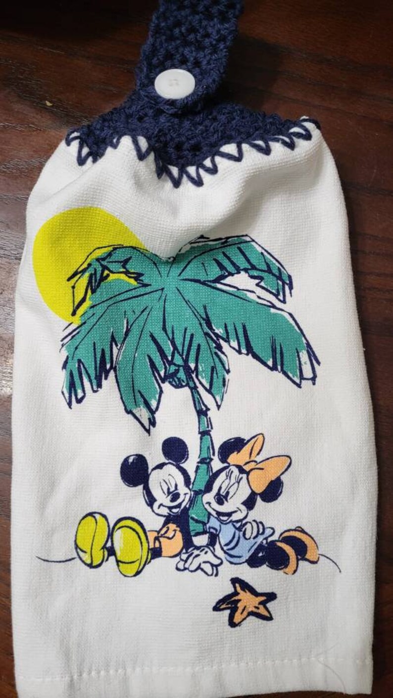 May include: White kitchen towel with a blue crocheted hanging loop and a button. The towel features a cartoon image of Mickey Mouse and Minnie Mouse under a palm tree with a yellow sun.