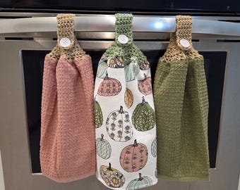May include: Three hanging kitchen towels. The first is a solid pink, the second has a white background with pumpkin illustrations, and the third is a solid green. Each towel has a crocheted top with a button.