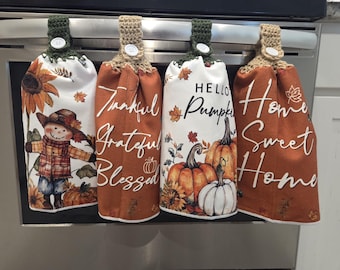 May include: Four decorative kitchen towels with crochet tops hang from a metal bar. The towels feature fall-themed designs, including a scarecrow, pumpkins, sunflowers, and the phrases "Thankful, Grateful, Blessed" and "Hello Pumpkin."